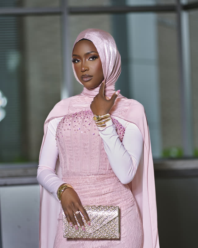 Portrait of a woman in a blush-pink hijab and beaded gown holding a gold clutch in evening light