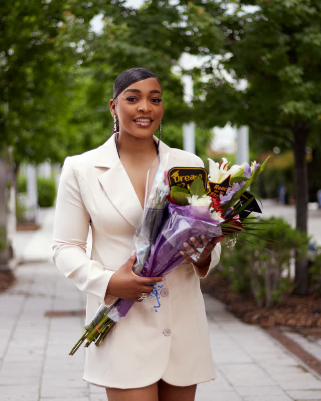 Outdoor graduation portrait of a woman in a white blazer dress holding a wrapped bouquet on a tree-lined walkway