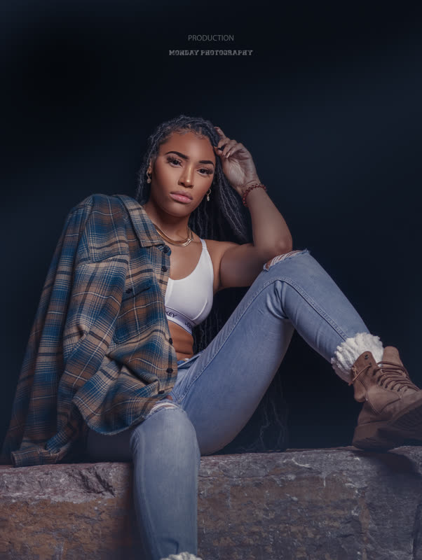 Editorial studio portrait of a woman in an open flannel shirt and ripped jeans seated against a dark backdrop