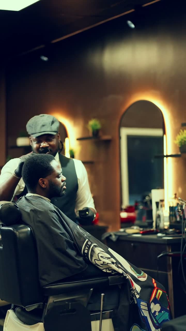 Film still: a barber in a flat cap and vest standing behind a client seated in a warm, wood-toned barbershop