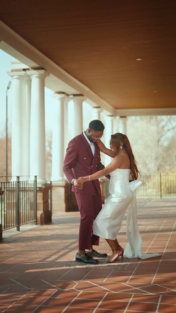 Film still: a couple dancing under a colonnade, the bride in a white gown and red heels, the groom in a burgundy suit
