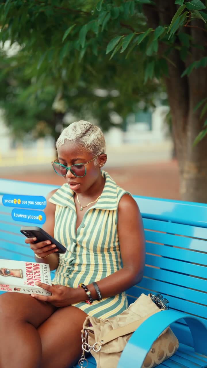 Film still: a woman in a green-striped dress and blue sunglasses seated on a blue bench, reading