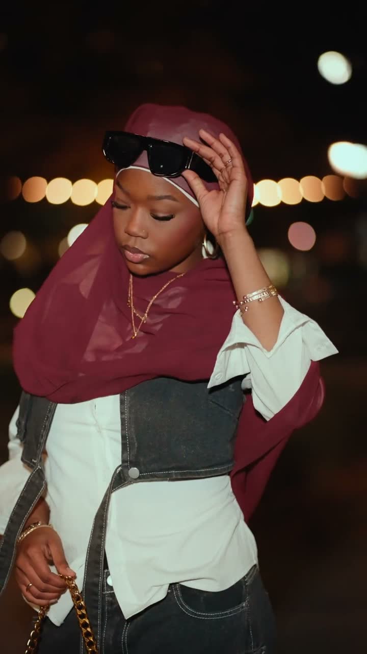 Film still: a woman in a burgundy hijab and denim adjusting her sunglasses against blurred night city lights