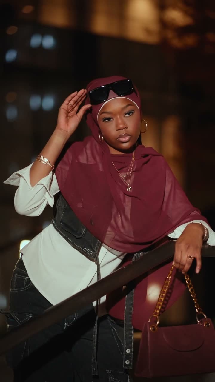 Film still: a woman in a burgundy hijab leaning on a railing with a chain bag under night city lights
